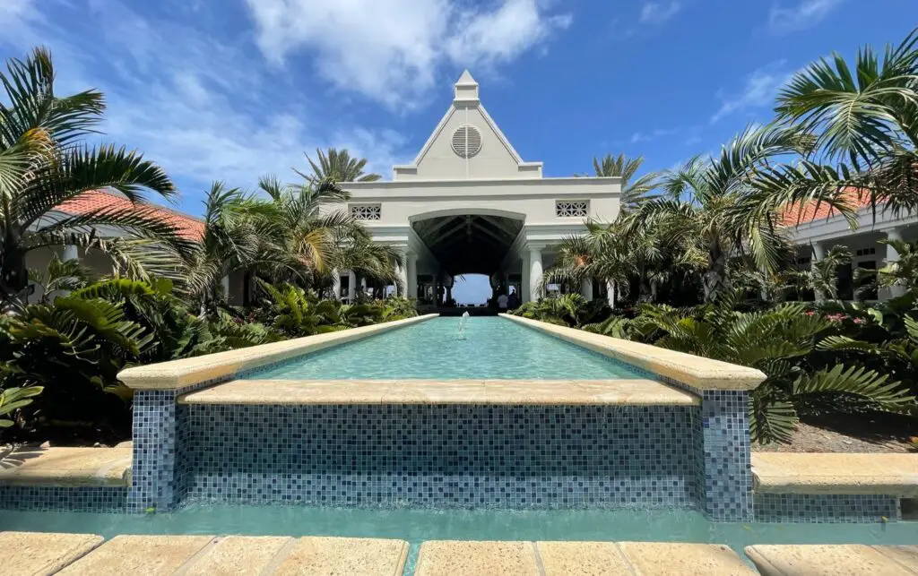 The view of the entrance of the Hotel Marriott A long pool surrounded by palm trees and green plants and the lobby in the background under a bright blue sky.- Curacao Marriott Beach Resort- Resort - all inclusive resorts at curacao Willemstad Curacao. Island of Curaçao - exploringcuracao.com